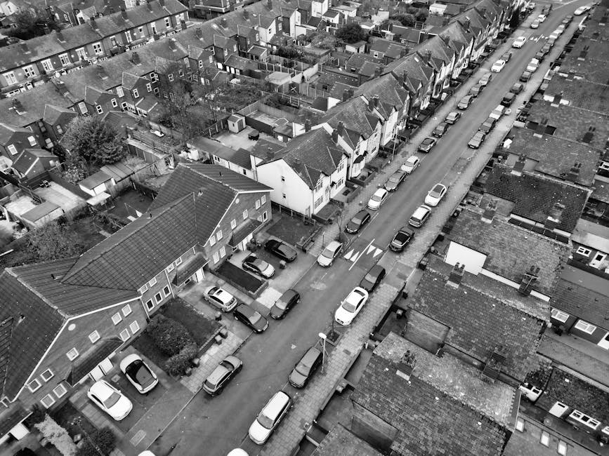 A black-and-white aerial photograph of a residential street showing closely spaced houses and terraced properties. The street is lined with parked cars on each side, including sedans and hatchbacks, w
